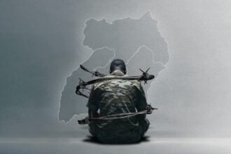 A man in camouflage uniform sits on the floor with barbed wire wrapped tightly around him. Behind him is a ghostly outline of Uganda’s map, symbolizing national repression and military entrapment.